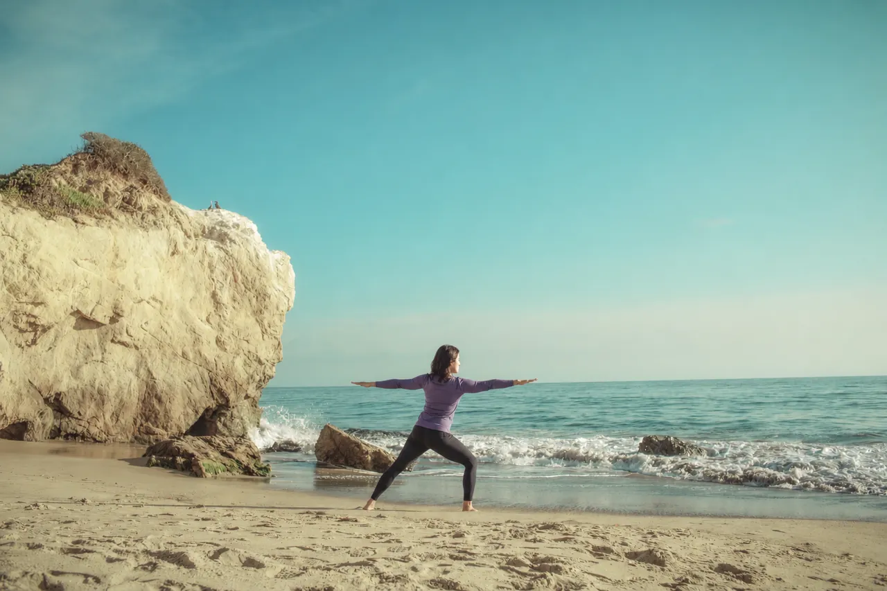 A woman in a purple top in a yoga pose beside a coastal cliff and pale blue sea