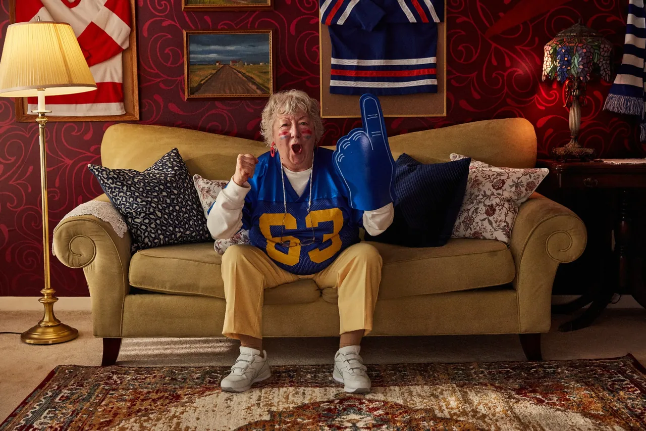 A cheering sports fan in a blue jersey raising a foam finger on a gold sofa